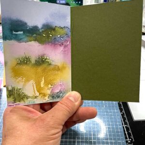 A hand holds an open green card with a landscape scene on the inside.