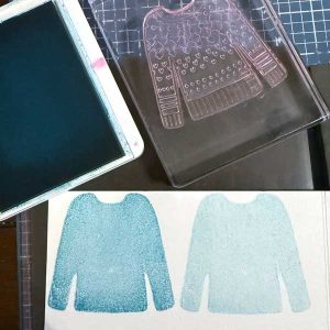 A blue ink pad lies next to a clear stamp shaped like a sweater. The solid image of a sweater is stamped onto white paper twice, one lighter than the other.